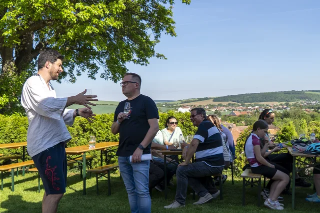 Hosté se sklenkami u stolů na zahradě, za živým plotem výhled na Sady a kopce
