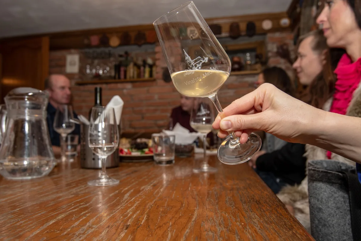 Ruka zvedá sklenku bílého vína s logem Vinařství Medek, za ní hosté u dřevěného stolu ve sklepě