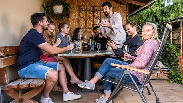 Posezení pod pergolou: společnost u kulatého kamenného stolu s lahvemi a sklenkami, kolem dřevěná lavice a zahradní křesla
