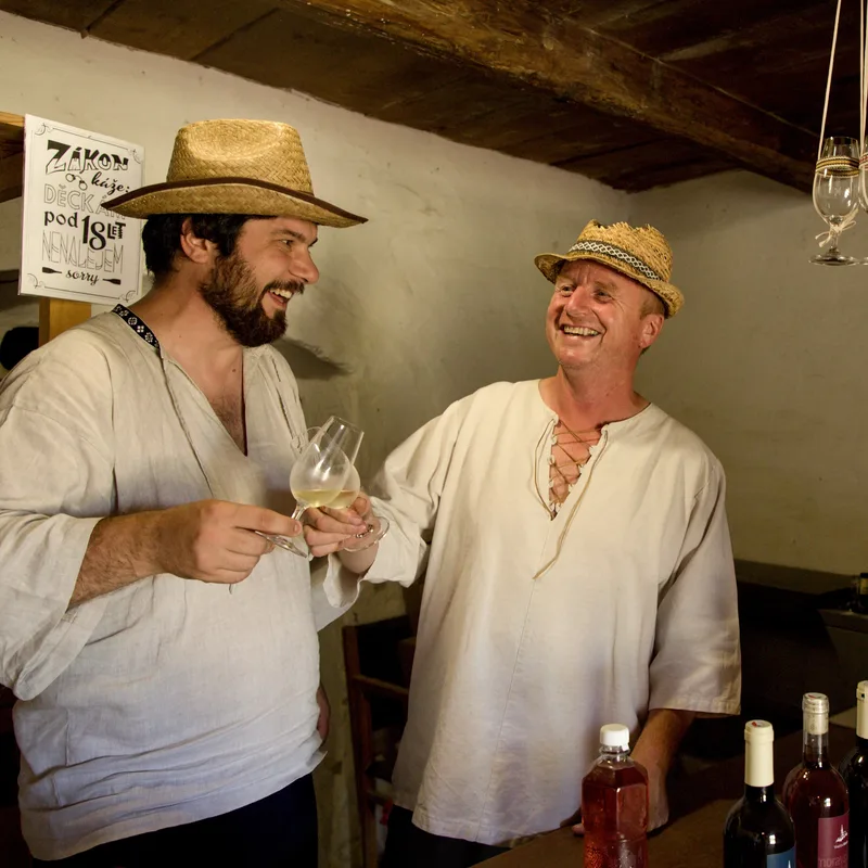 Jožka (vpravo) a Jaromír, oba v krojových košilích a slaměných kloboucích, přípíjejí si vínem; na pultu lahve vinařství, nad nimi zavěšené sklenice.