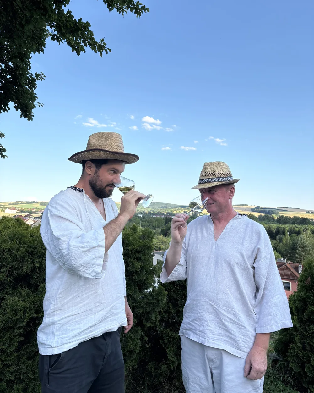 Vpravo vinař Jožka voní ke sklence bílého vína, vlevo jeho zeťák Jaromír. Oba v krojových košilích a slaměných kloboucích, za nimi kopce Slovácka.