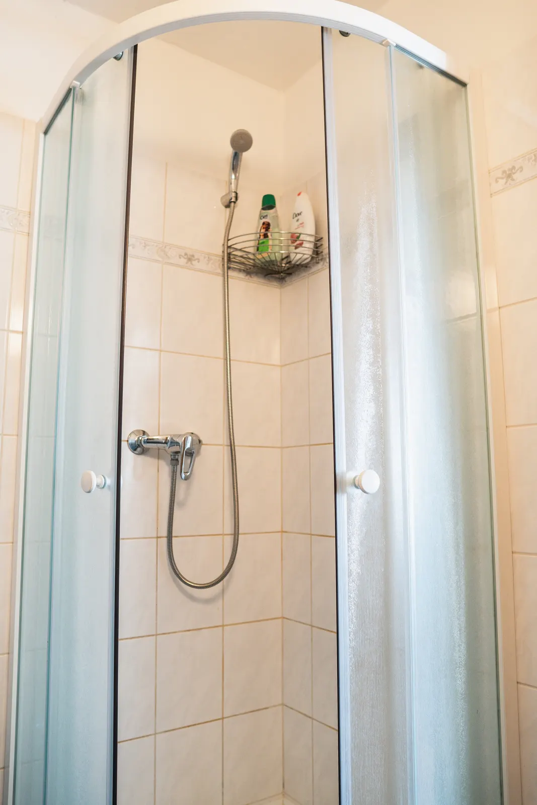 Koupelna Apartmánu: zaoblený sprchový kout s posuvnými dveřmi v bílém obkladu