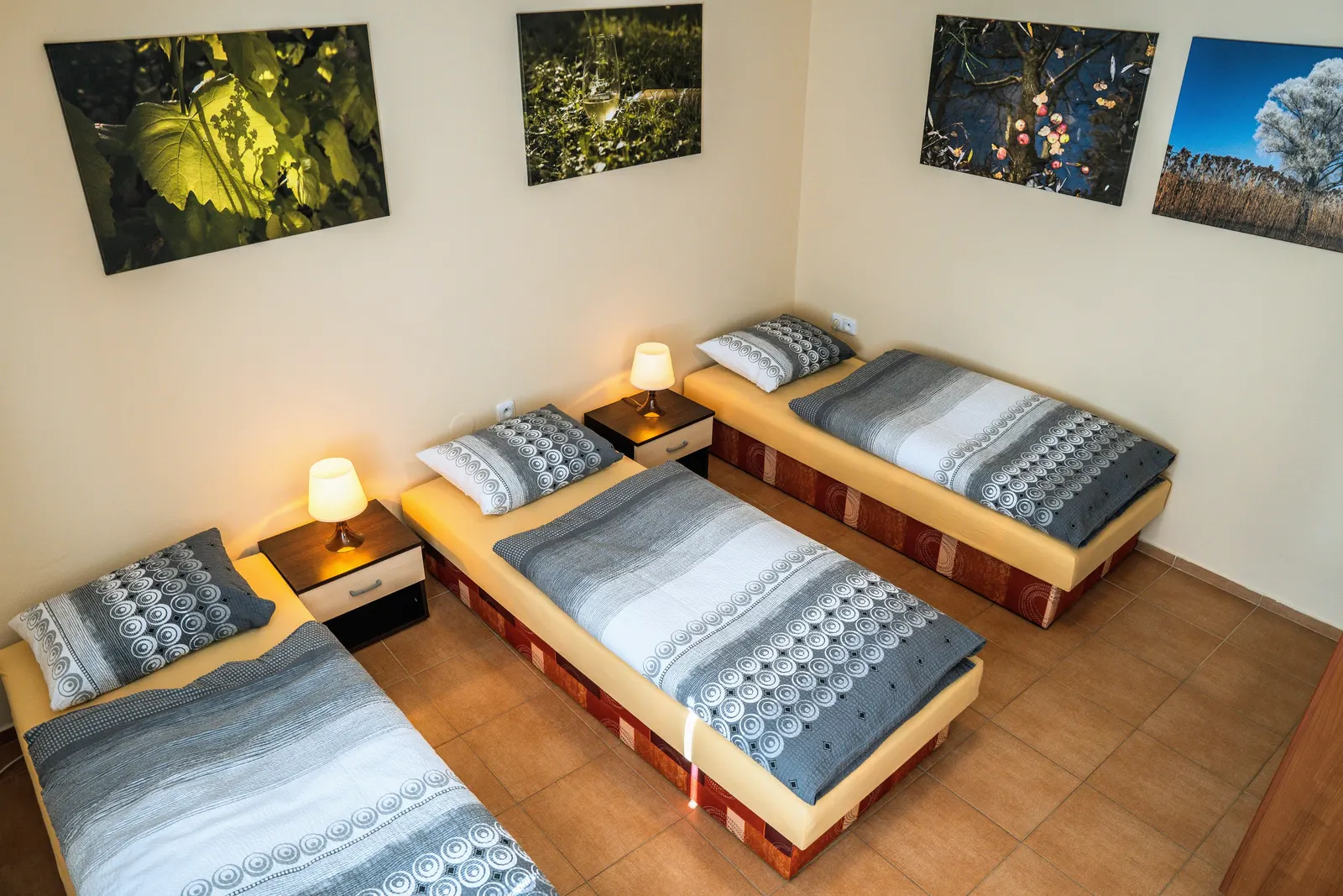Druhá ložnice Apartmánu: tři jednolůžkové postele podél zdí, noční stolky s lampičkami a fotoobrazy z vinohradu a sadu