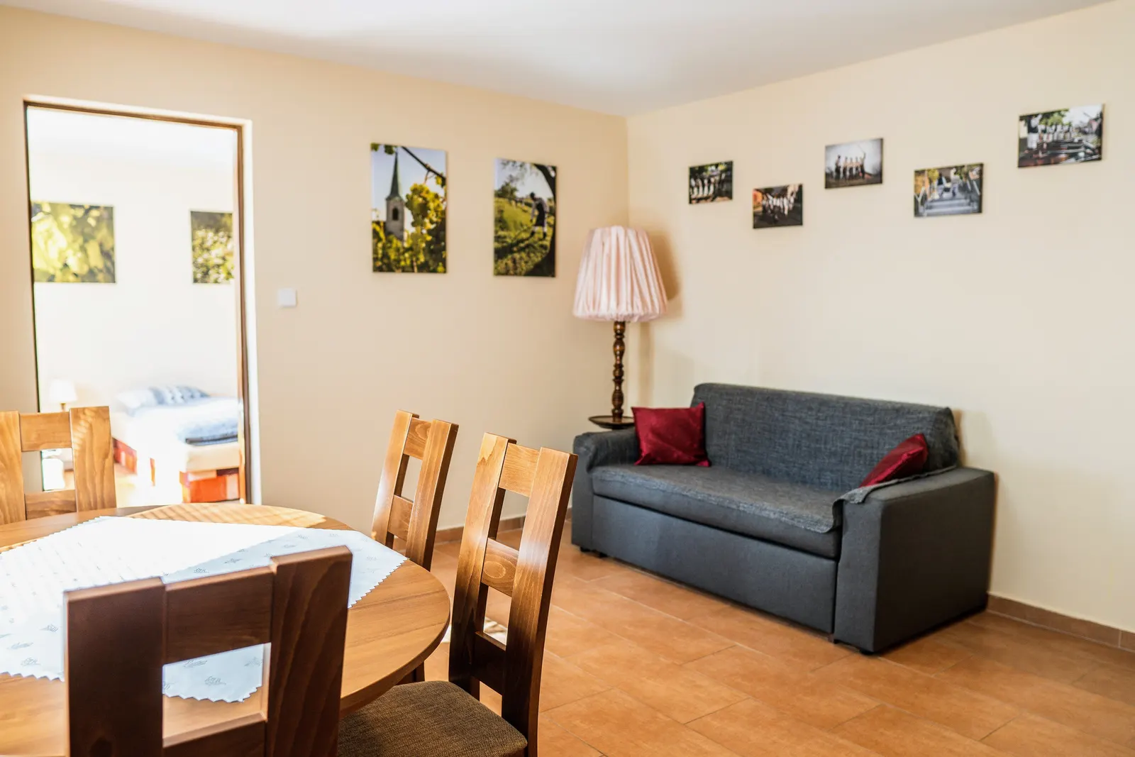 Obývací pokoj Apartmánu: kulatý jídelní stůl se čtyřmi židlemi, šedá pohovka s vínovými polštáři, stojací lampa a otevřenými dveřmi průhled do ložnice