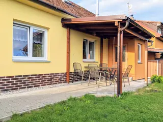 Apartmán zvenku: vlastní vchod pod krytou terasou se stolem a kovovými židlemi, před ní zámková dlažba a trávník