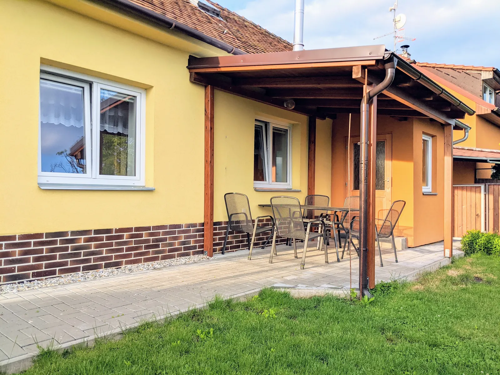Apartmán zvenku: vlastní vchod pod krytou terasou se stolem a kovovými židlemi, před ní zámková dlažba a trávník